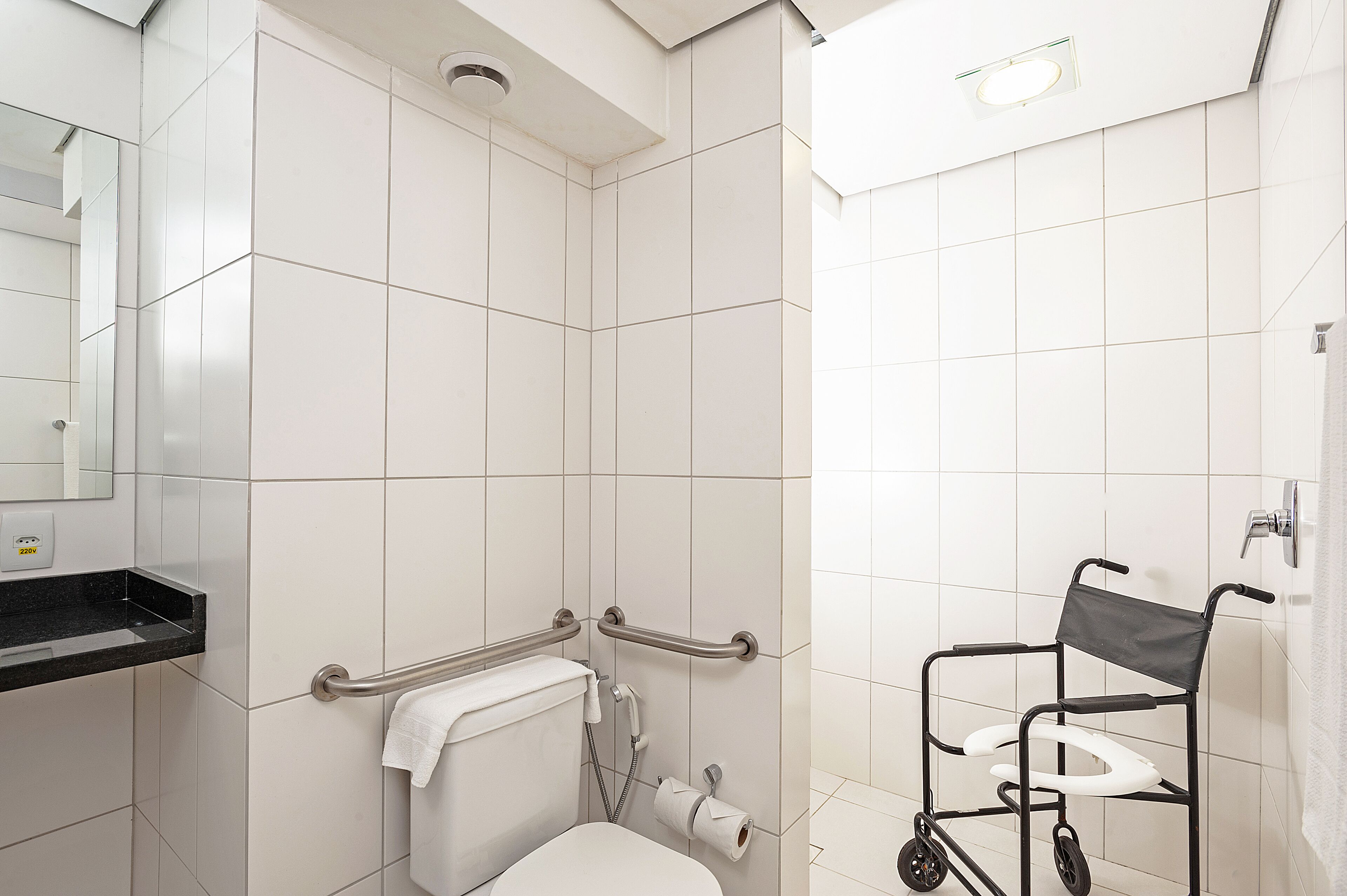 A bathroom with white tiles, a toilet, a handrail, a wheelchair, and a towel rack.