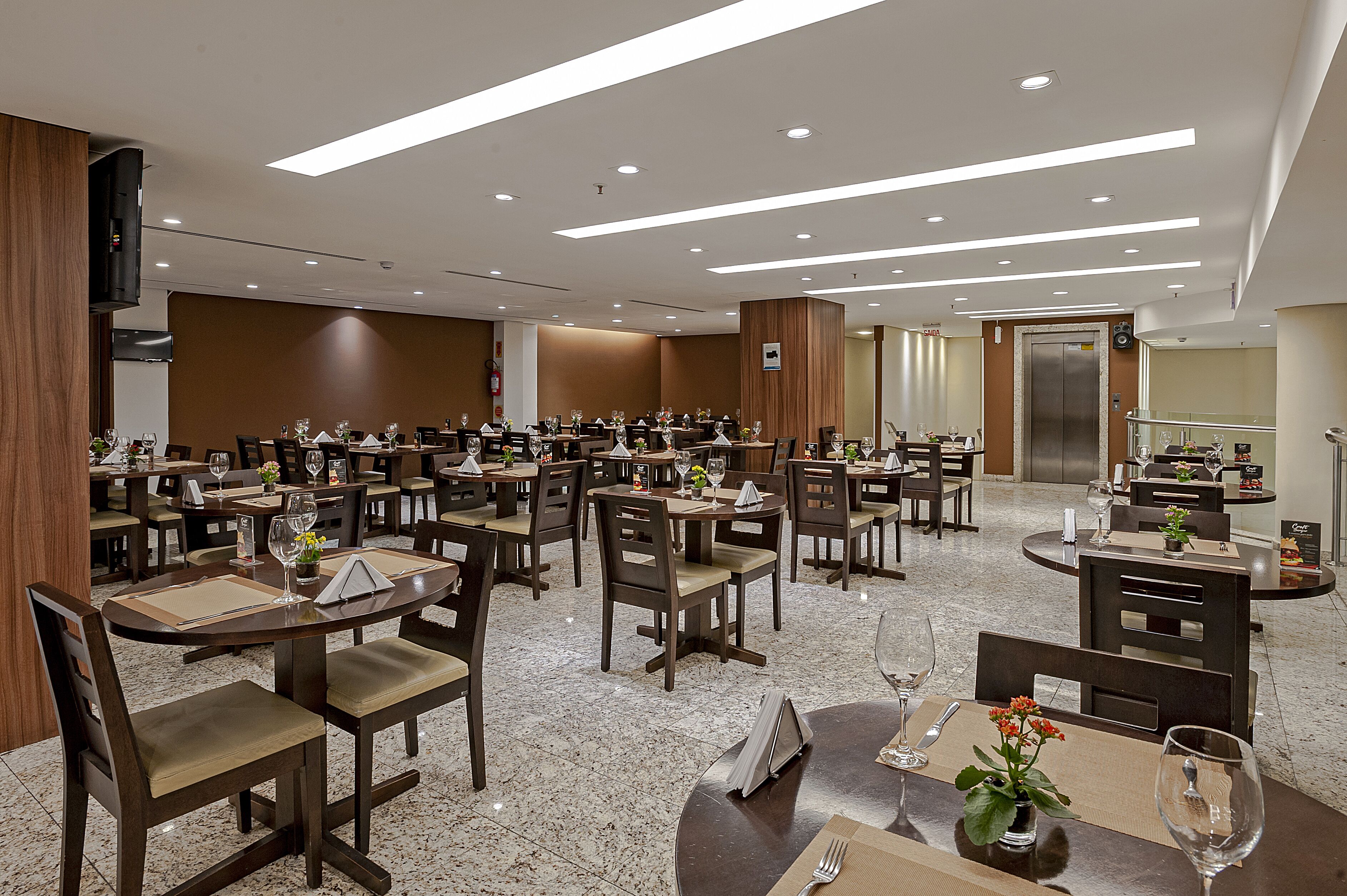 Ampla sala de jantar de hotel, com várias mesas e cadeiras, completa com louças preparadas e um design moderno no teto.