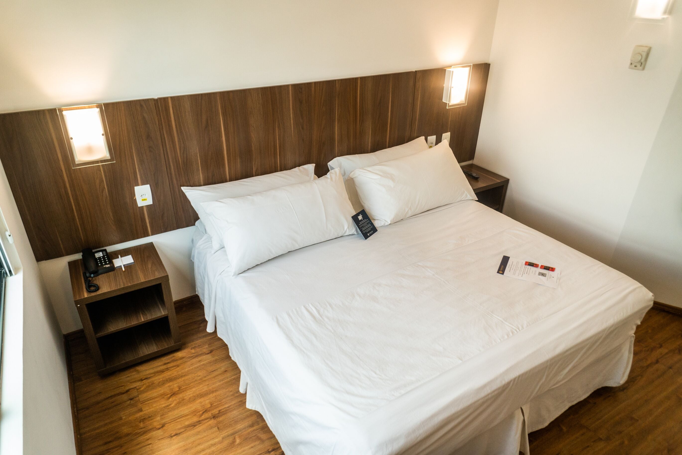 A hotel room with a wooden headboard, two bedside tables, a white bed with pillows, and a nightstand with a phone.