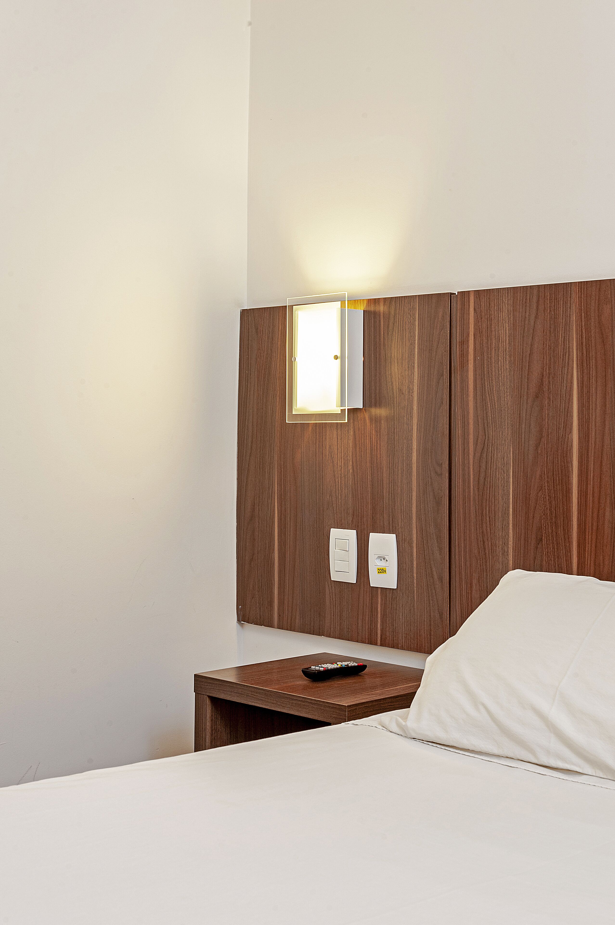 Una habitación de hotel con cabecera de madera, una mesita de noche con control remoto y una cama blanca.