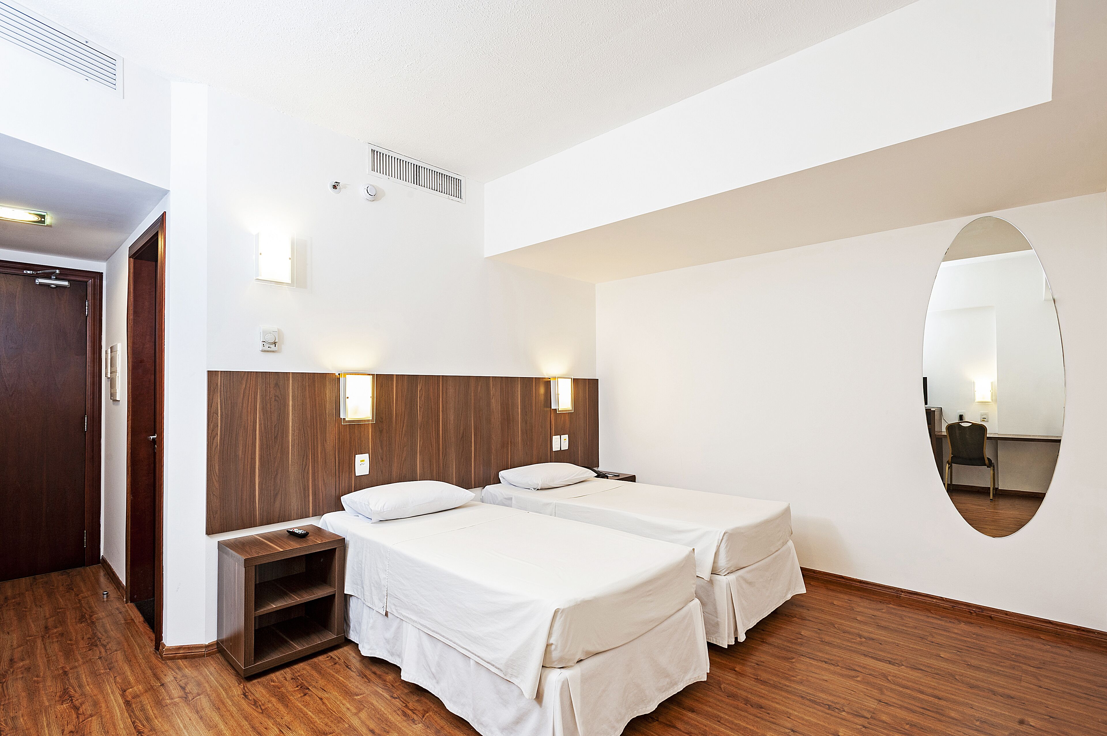 A hotel room with two single beds, a wooden headboard, a small bedside table, a mirror, and a wooden floor.