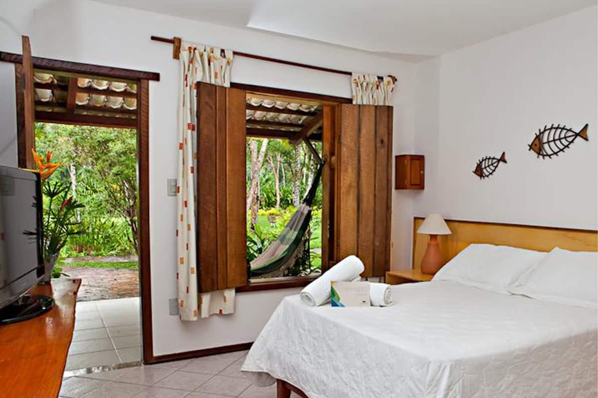 A hotel room with a bed, a television, a window with curtains, a hammock, and a view of greenery outside.