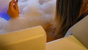 A person relaxing in a bathtub filled with bubbles, holding a glass of white wine, with a scenic view of mountains through a window. - Villa Bella Hotel & SPA Gramado (Gramado)