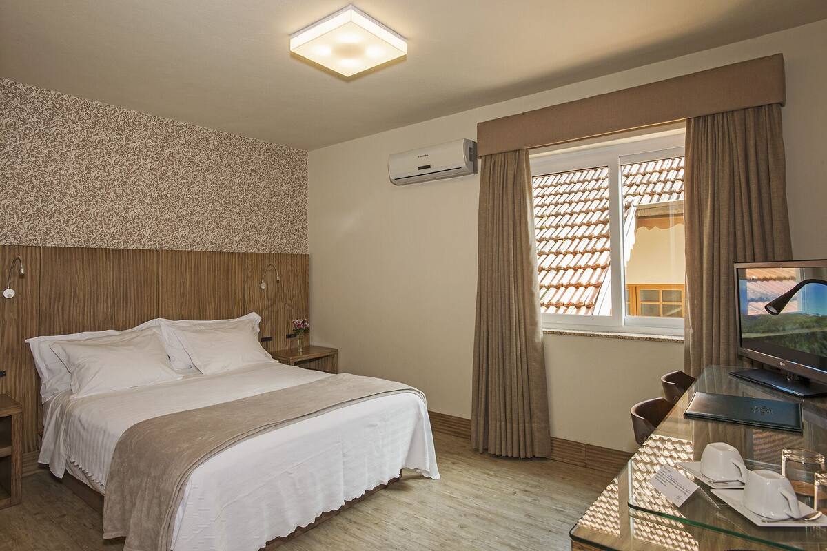 A hotel room with a bed, a TV, a desk with a glass top, and a window with curtains.