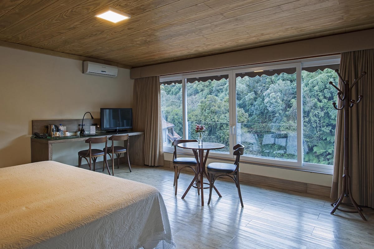 A hotel room with a bed, a desk with a TV, a chair, a table with chairs, and a large window with a view of trees.