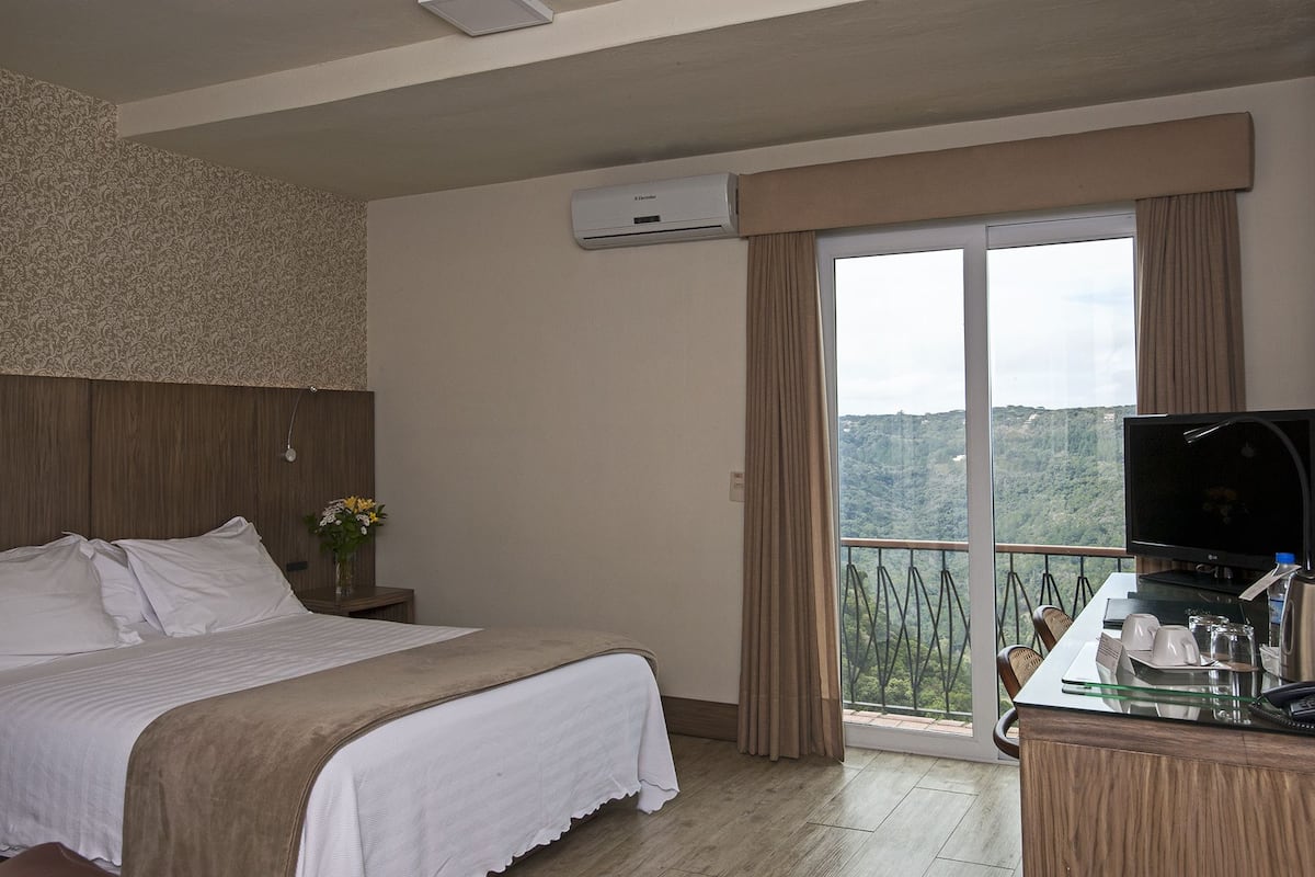 A hotel room with a bed, a desk with a TV, a window with curtains, and a view of a mountainous landscape.