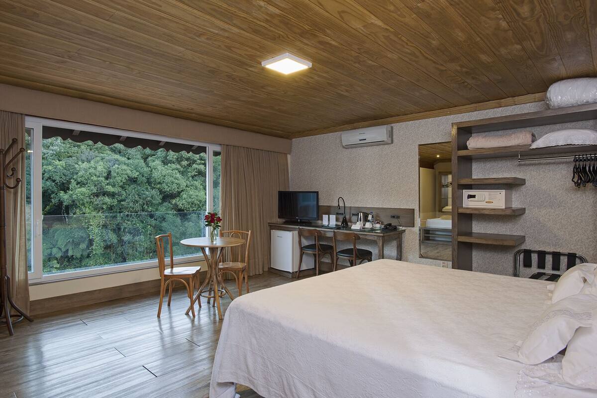 A hotel room with a bed, a desk with a computer, a chair, a small table with flowers, and a large window with a view of trees.