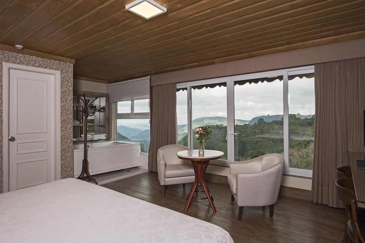 A hotel room with a large bed, two armchairs, a small table with a vase, a television, and a view of the mountains.
