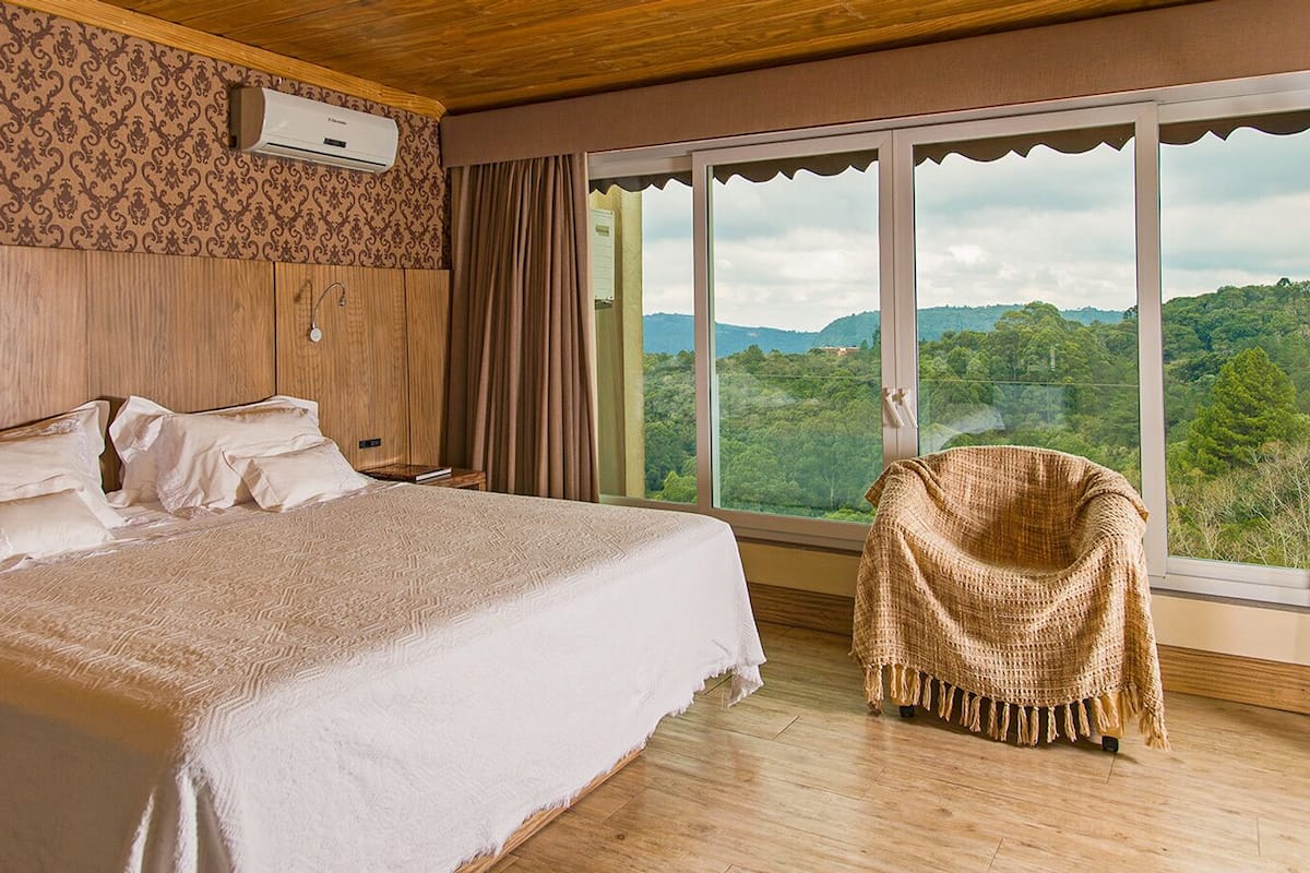 A bedroom with a large bed, a wooden headboard, a window with a view of the forest, and a chair with a blanket.