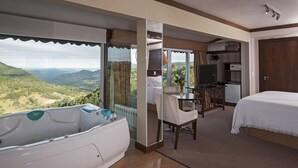 A hotel room with a large bathtub, a bed, a desk with a TV, and a view of the mountains. - Villa Bella Hotel & SPA Gramado (Gramado)
