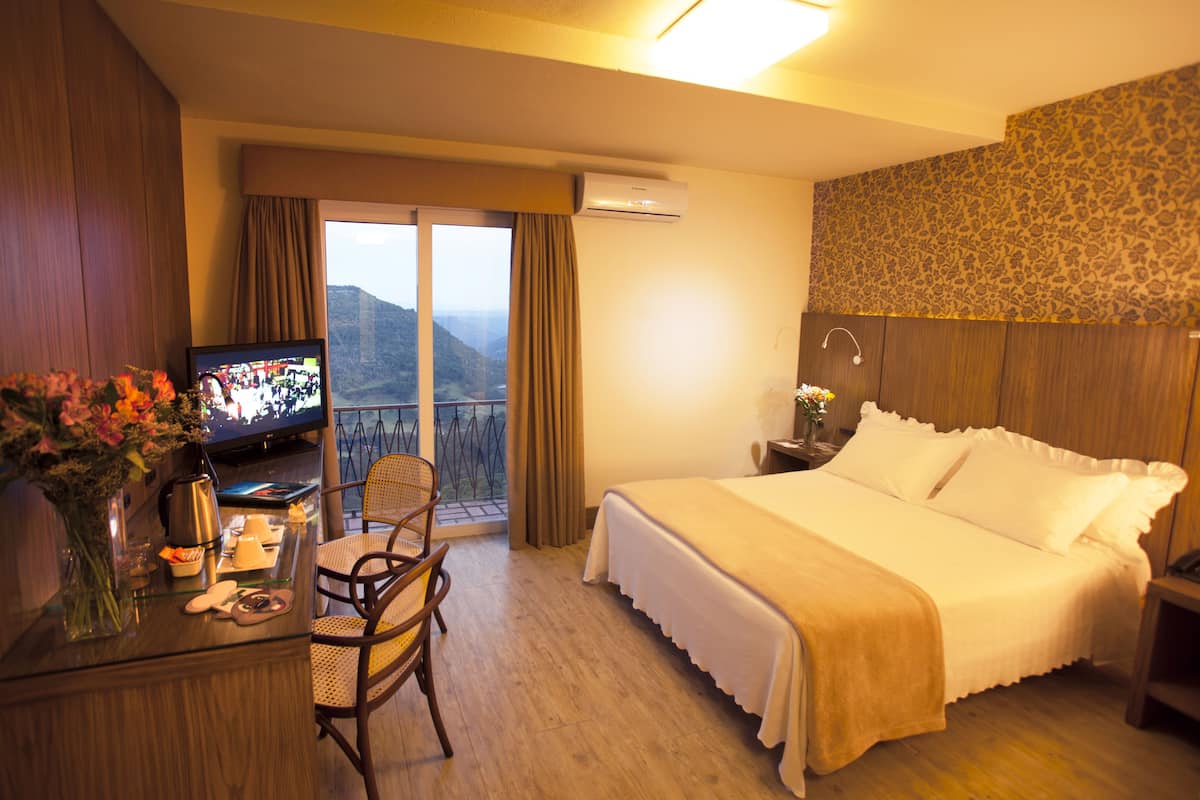A hotel room with a bed, a desk with a television, a chair, and a view of mountains through a balcony.