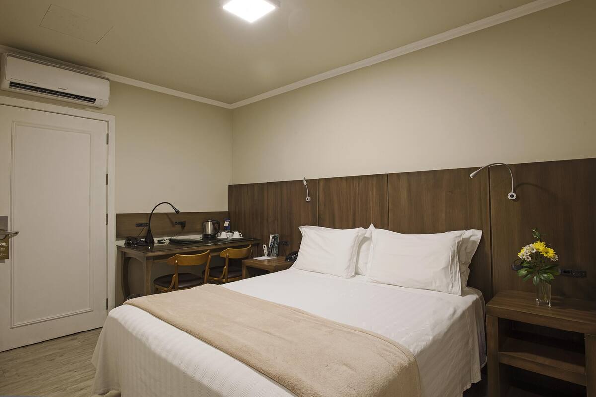 A hotel room with a bed, a desk, a chair, a nightstand, and a wall-mounted air conditioner.