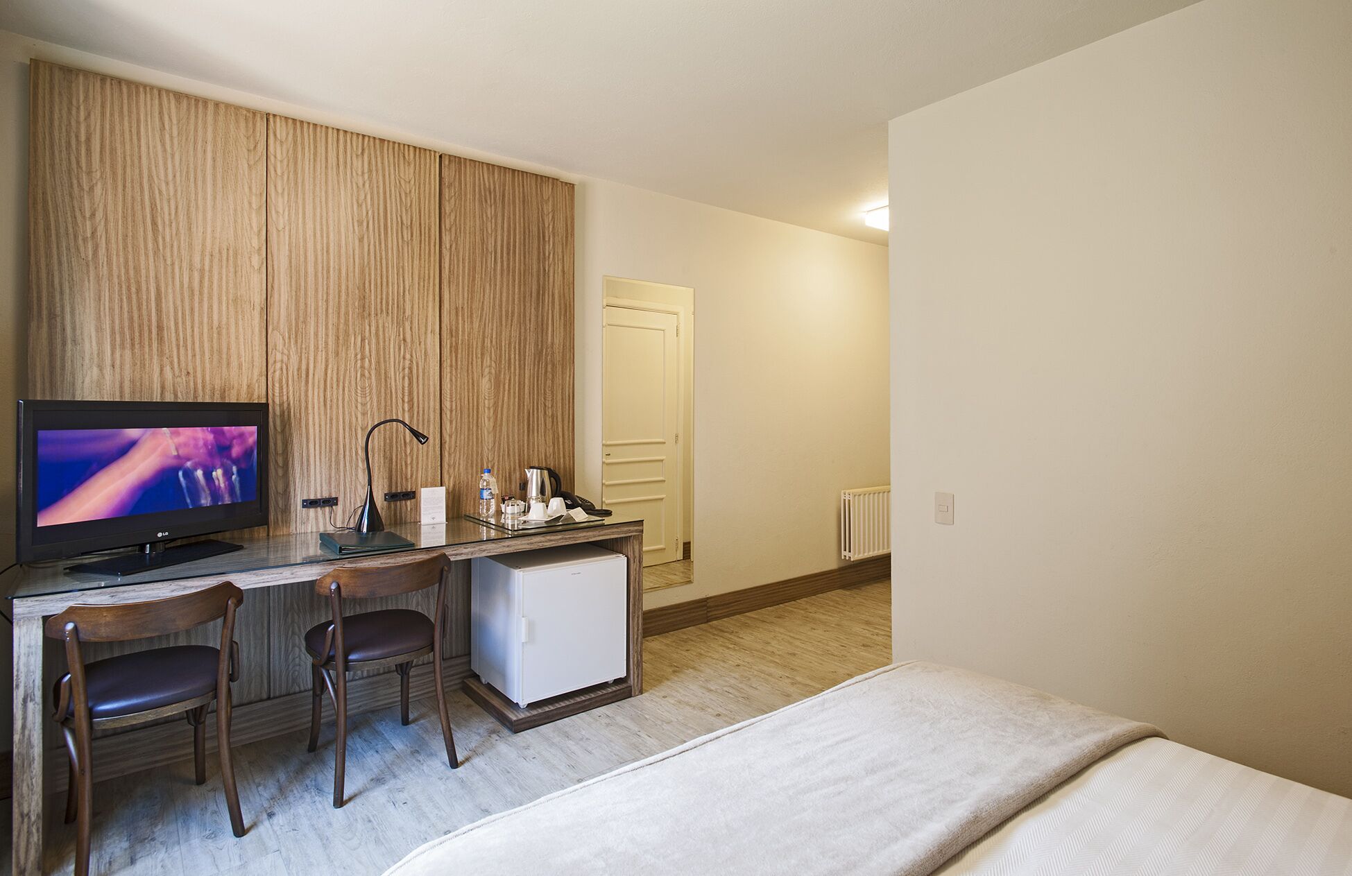 Ein Hotelzimmer mit Bett, Schreibtisch mit Fernseher, zwei Stühlen, kleinem Kühlschrank und einer Wandpaneele aus Holz.