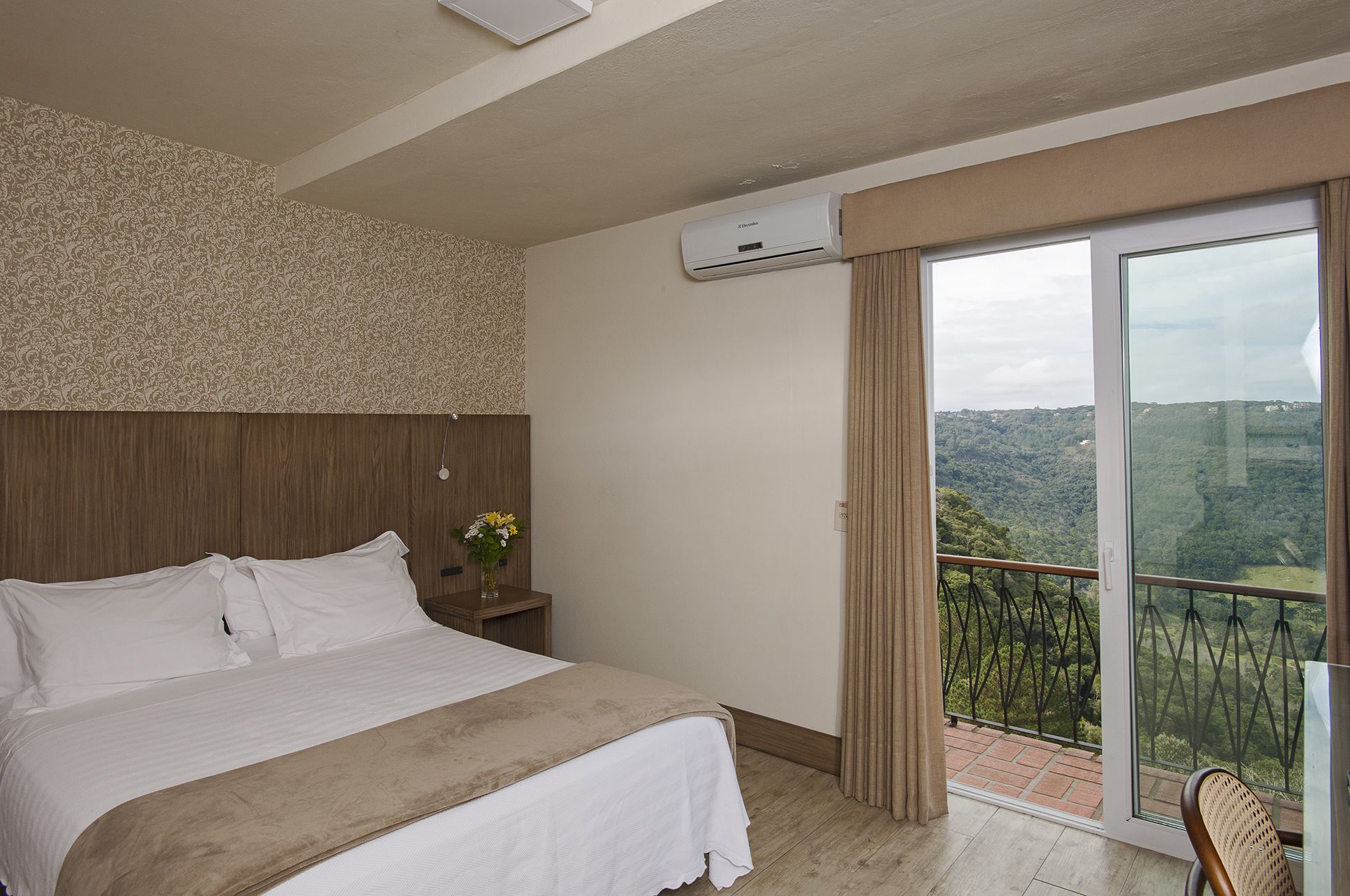 A bedroom with a bed, a nightstand, a vase with flowers, an air conditioning unit, and a balcony with a view of the countryside.