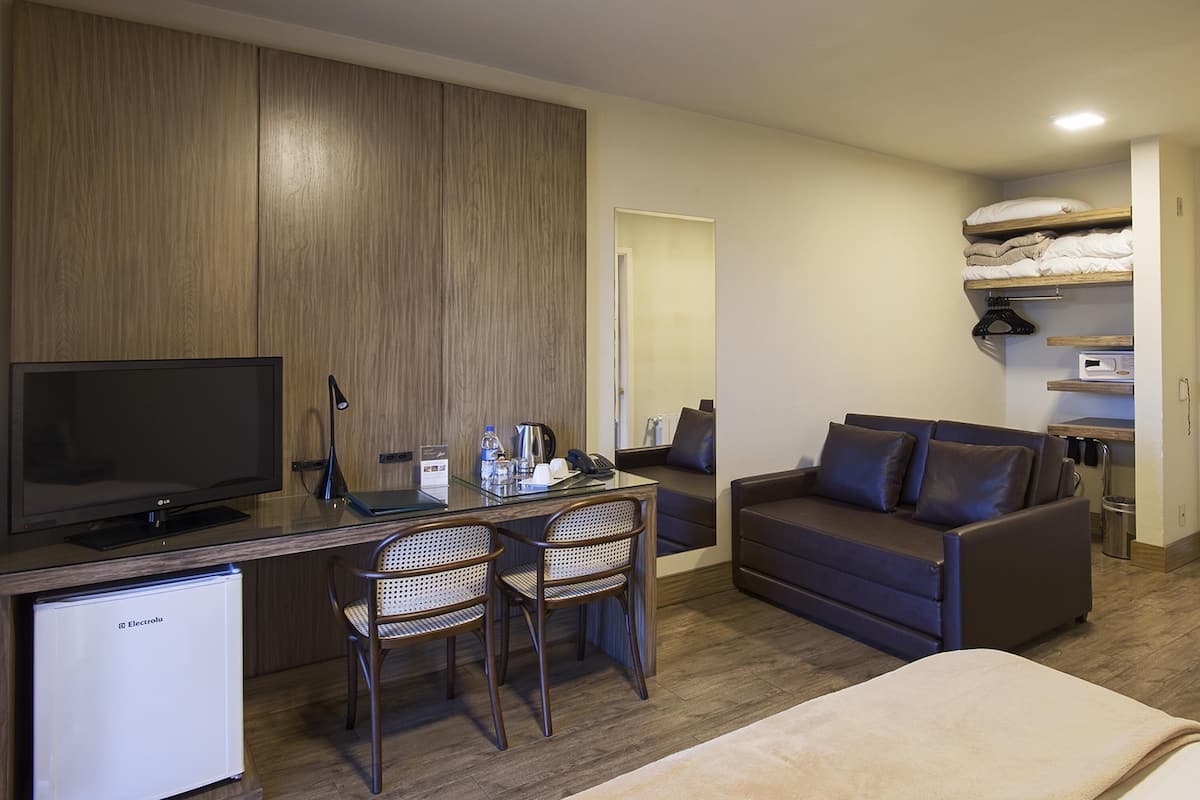 A compact hotel room with a flat-screen TV, a small fridge, a desk with a lamp, two wicker chairs, a sofa, and a bunk bed with a hanging rack.