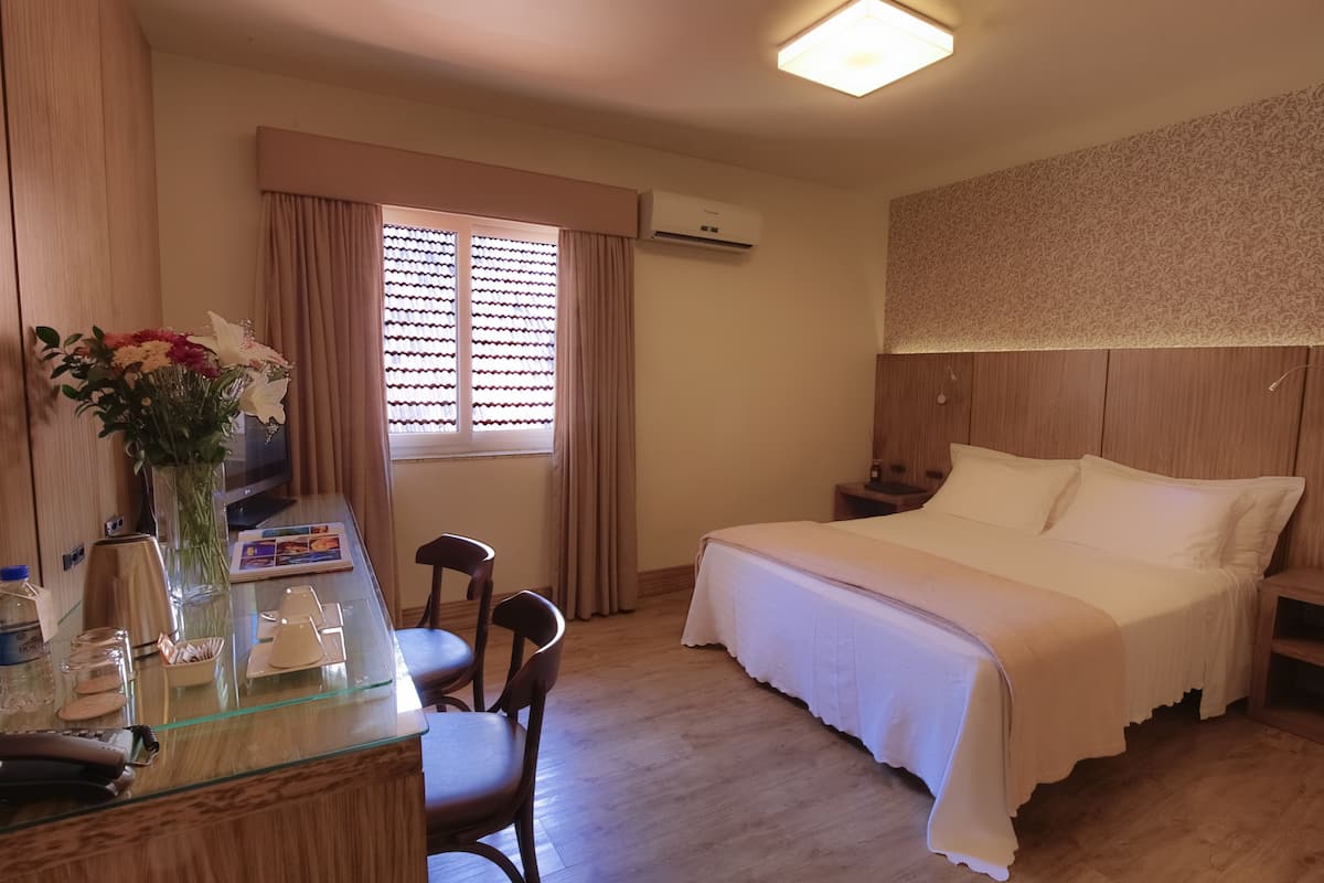 A hotel room with a bed, bedside table, chairs, a desk with a vase of flowers, and a window with curtains.