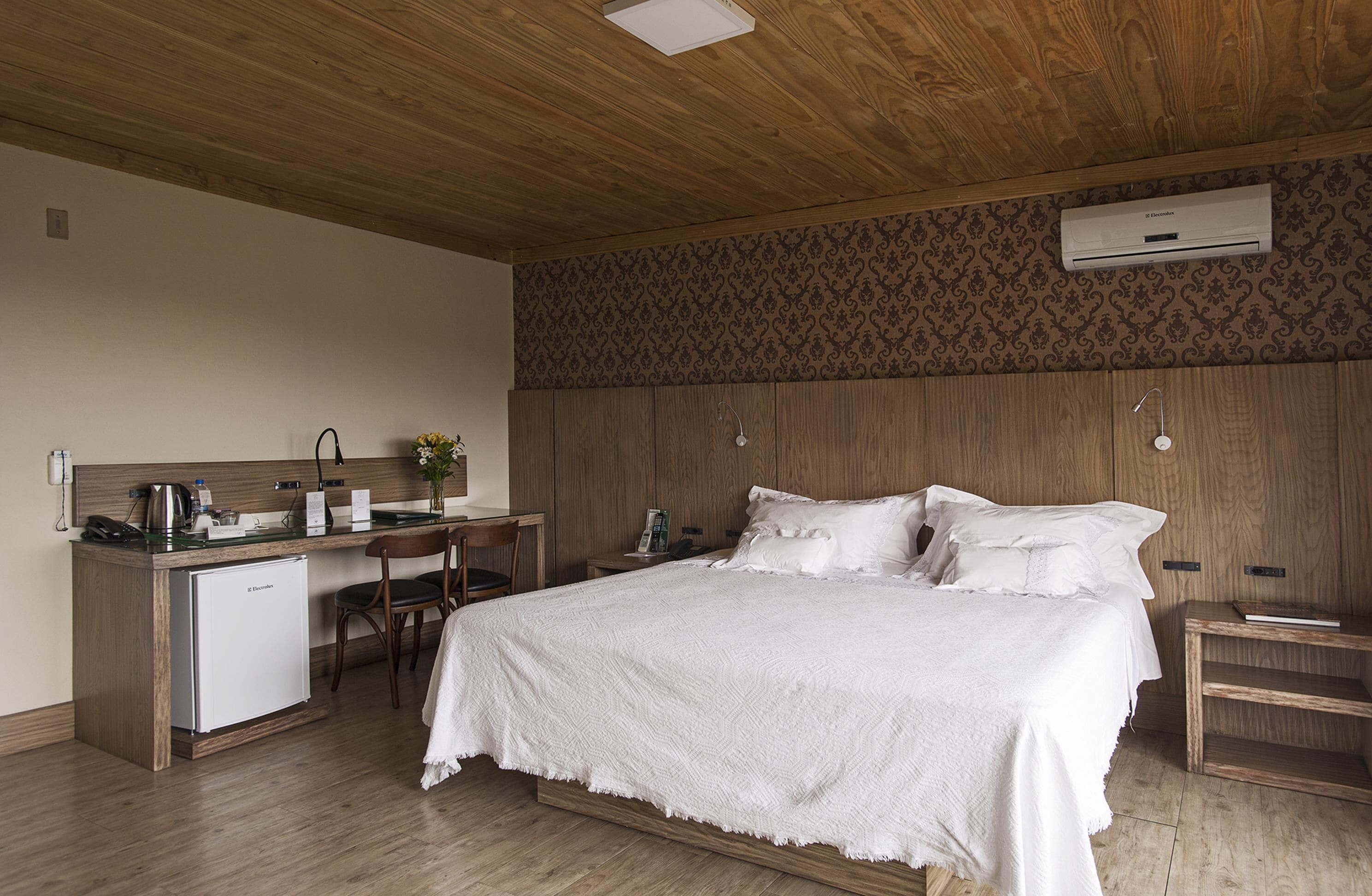Un dormitorio con techo de madera, una cama con ropa de cama blanca, un escritorio con una silla, un refrigerador pequeño y un equipo de aire acondicionado en la pared.