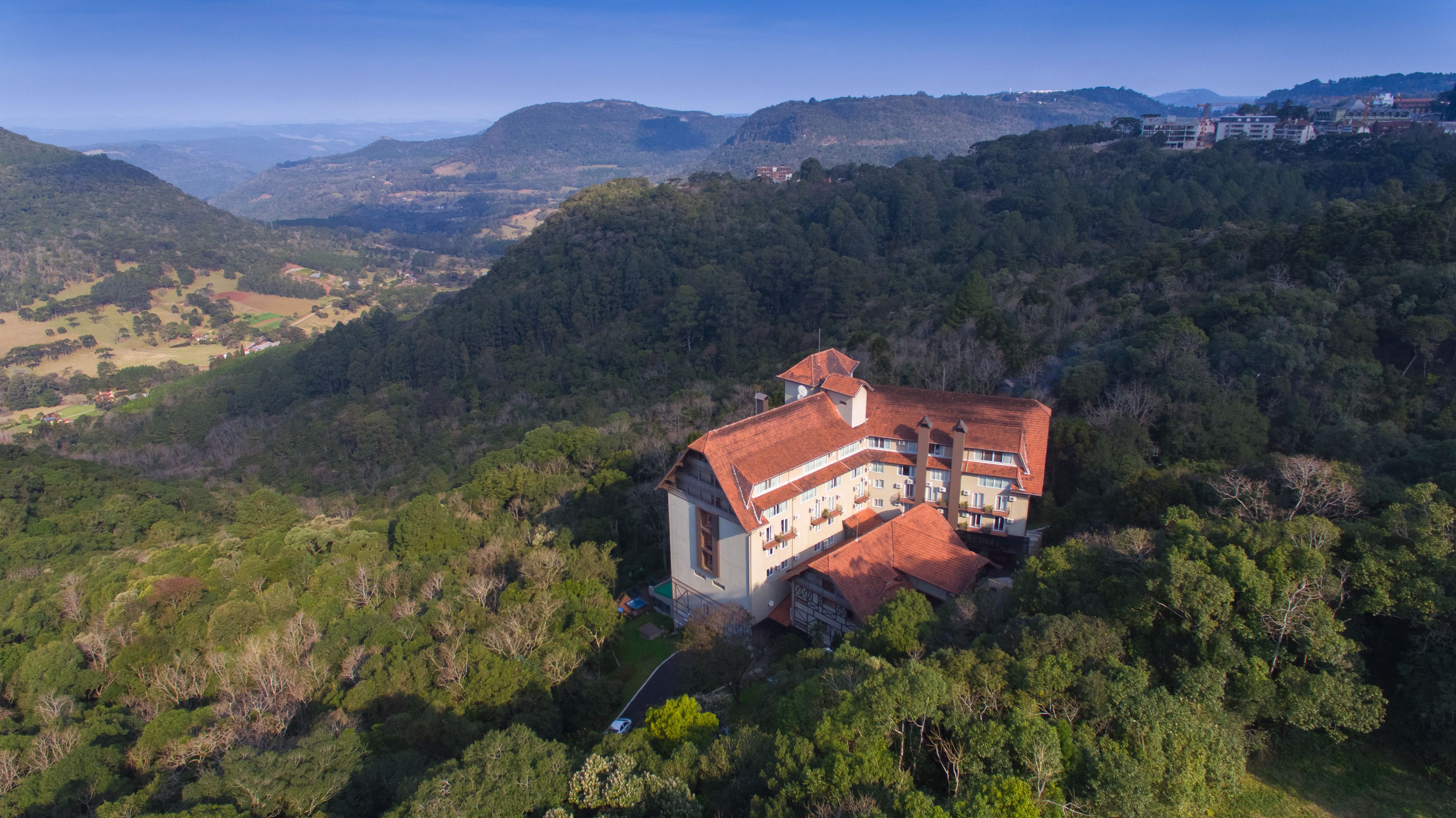 A large, multi-story building with a red-tiled roof, surrounded by dense forest, set in a hilly landscape.