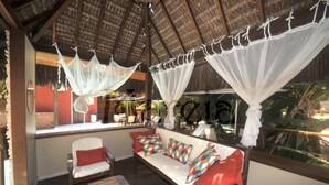 A covered outdoor seating area with a thatched roof, cushioned seating, and colorful cushions. - Pousada Mareia (Cairu)