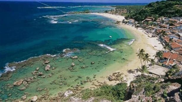 A coastal area with clear waters, a sandy beach, and a densely built-up area with buildings. - Pousada Mareia (Cairu)
