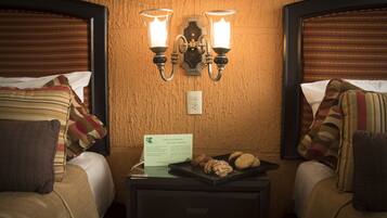 A hotel room with two beds, a nightstand with a tray of cookies, and a wall-mounted light fixture.