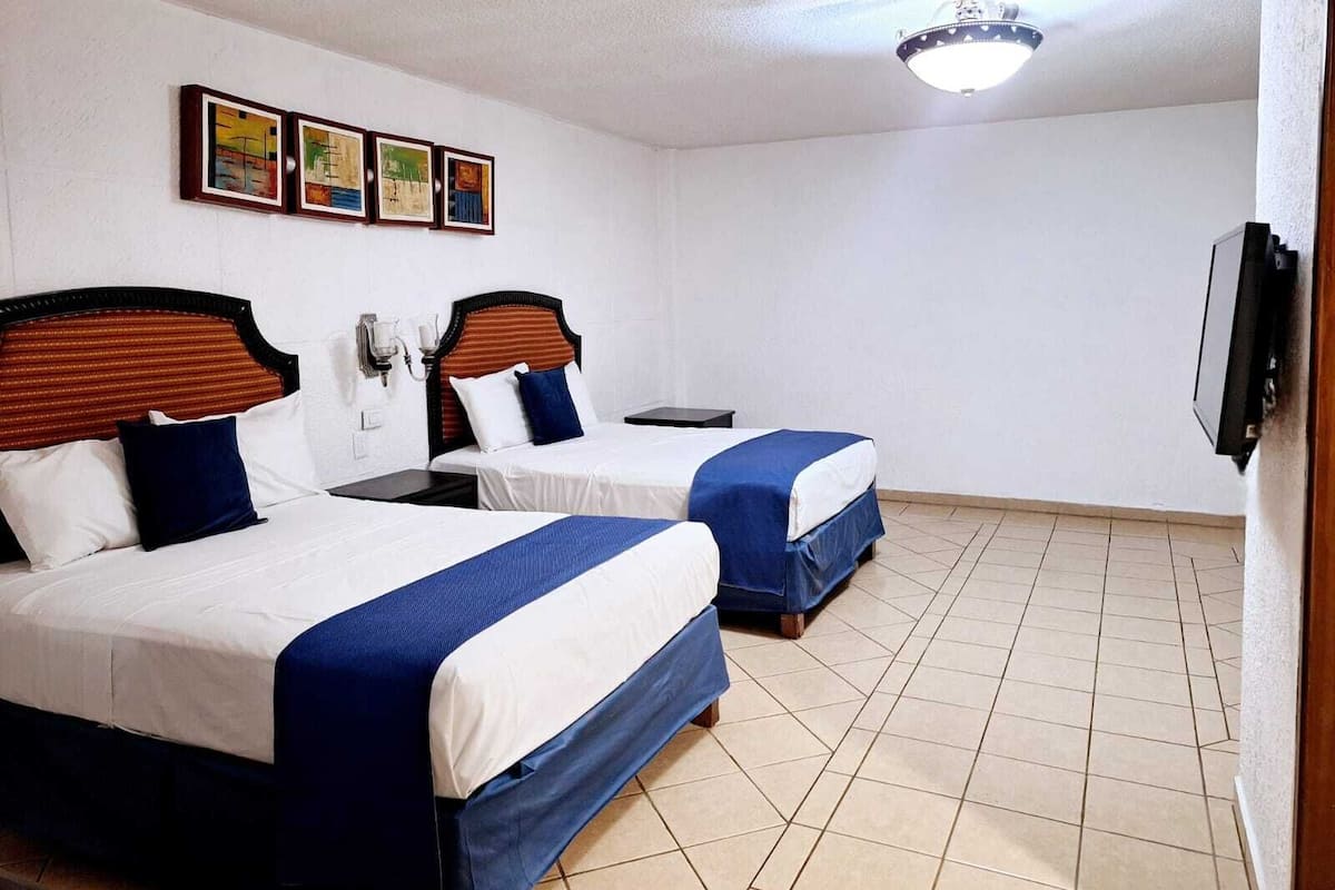A hotel room with two beds, each with a wooden headboard, white bedding, and blue accents. There are wall-mounted lights and a flat-screen TV on the wall.