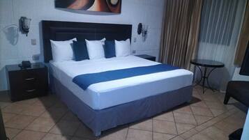 A hotel room with a large bed, a nightstand, a small table, and a wall-mounted lamp.
