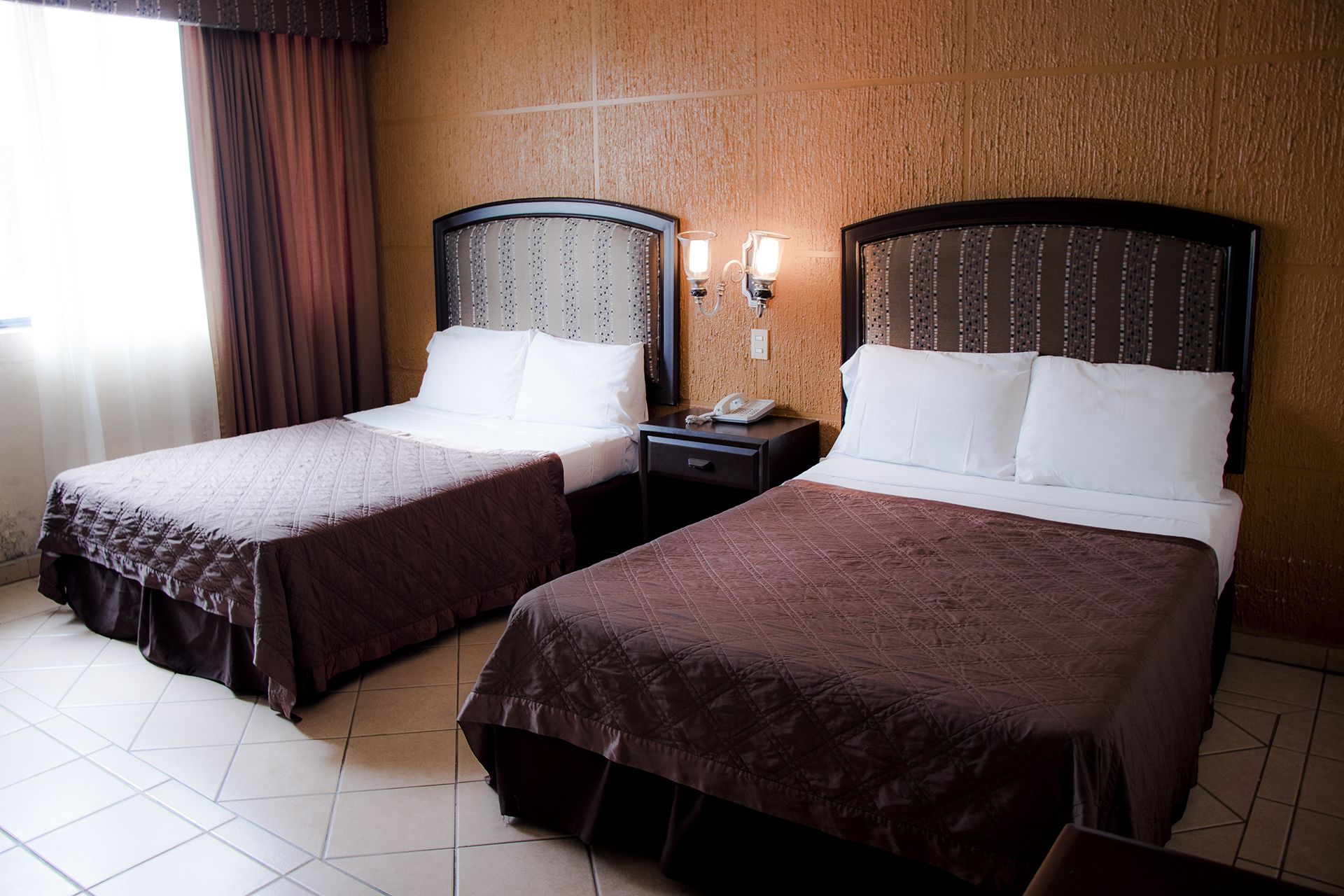A hotel room with two beds, each with a headboard and a nightstand with a phone.