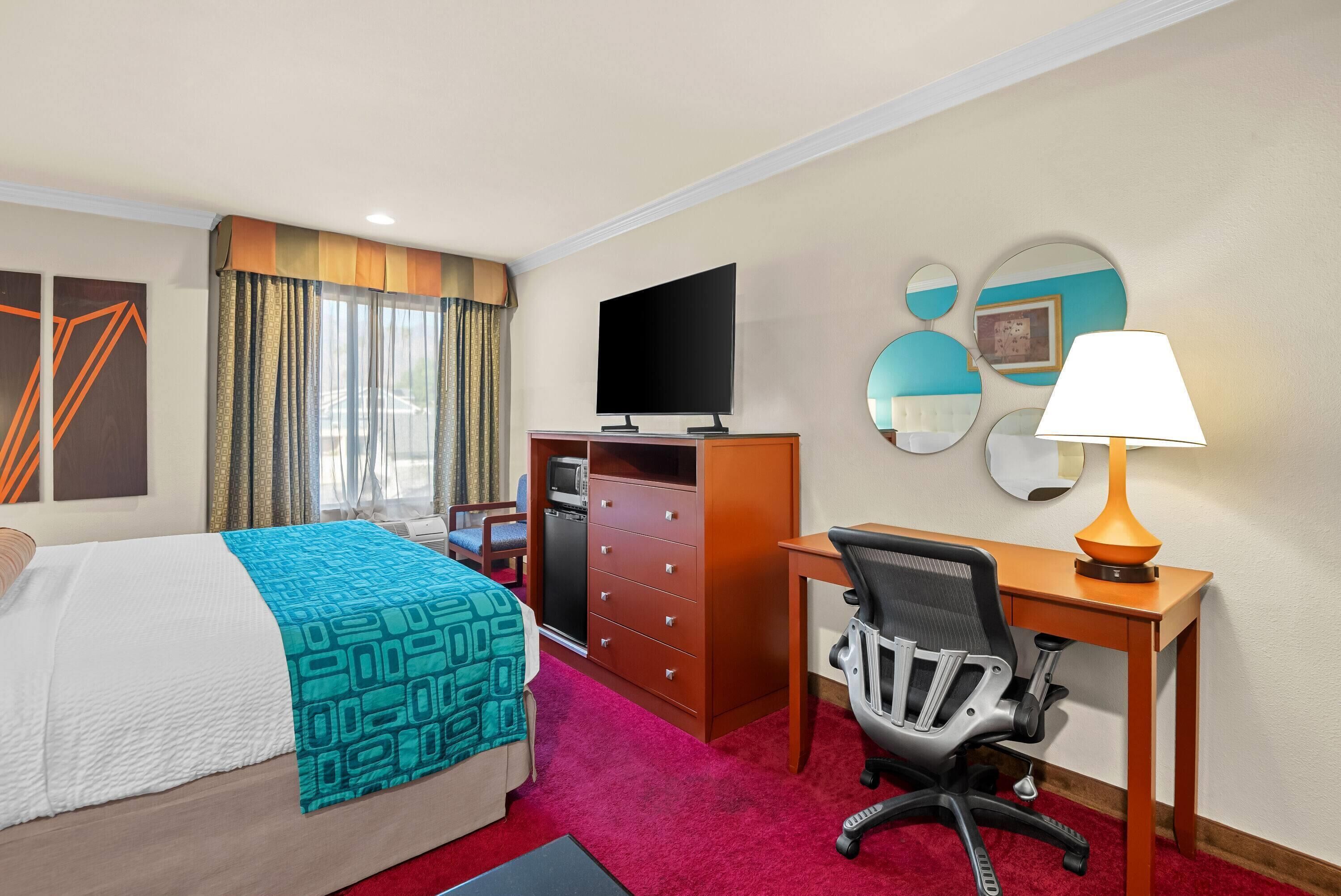 Habitación de hotel con cama, escritorio, silla, televisor y lámpara de escritorio.