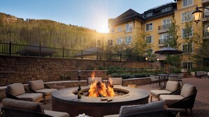 A circular fire pit surrounded by wicker chairs and tables, with a stone wall and a building in the background. - The Ritz-Carlton Club, Vail (Vail)