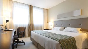 A hotel room with a bed, a desk with a laptop, a chair, a lamp, and a window with curtains.