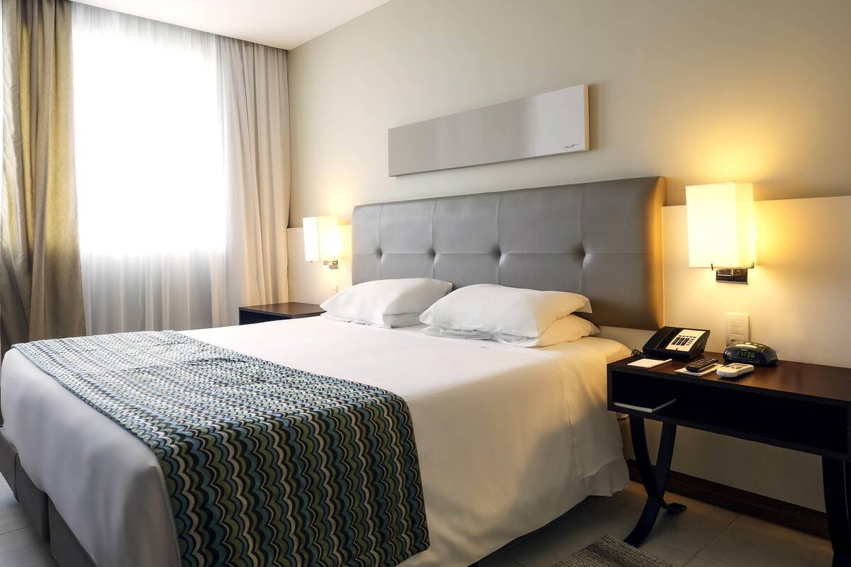 A hotel room with a large bed, two bedside tables, a nightstand, a lamp, a phone, and a clock.