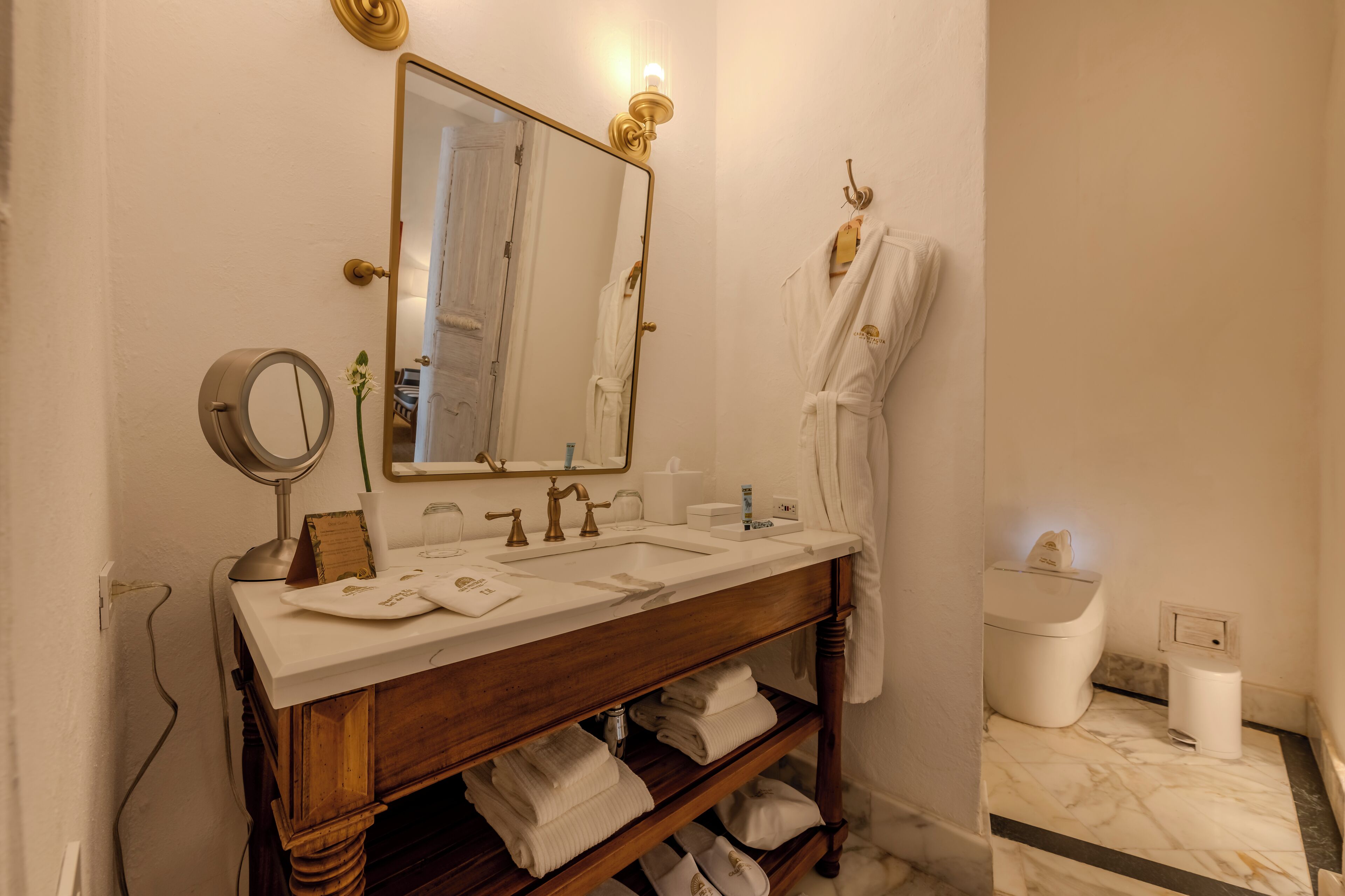 Ein Badezimmer mit einem Holzwaschtisch, einem großen Spiegel und einem weißen Waschbecken. Handtücher und ein Bademantel hängen an der Wand.