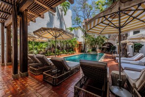 A poolside area with wicker lounge chairs and striped umbrellas, surrounded by lush greenery and a white building. - Casa Pestagua Relais Châteaux (Cartagena)
