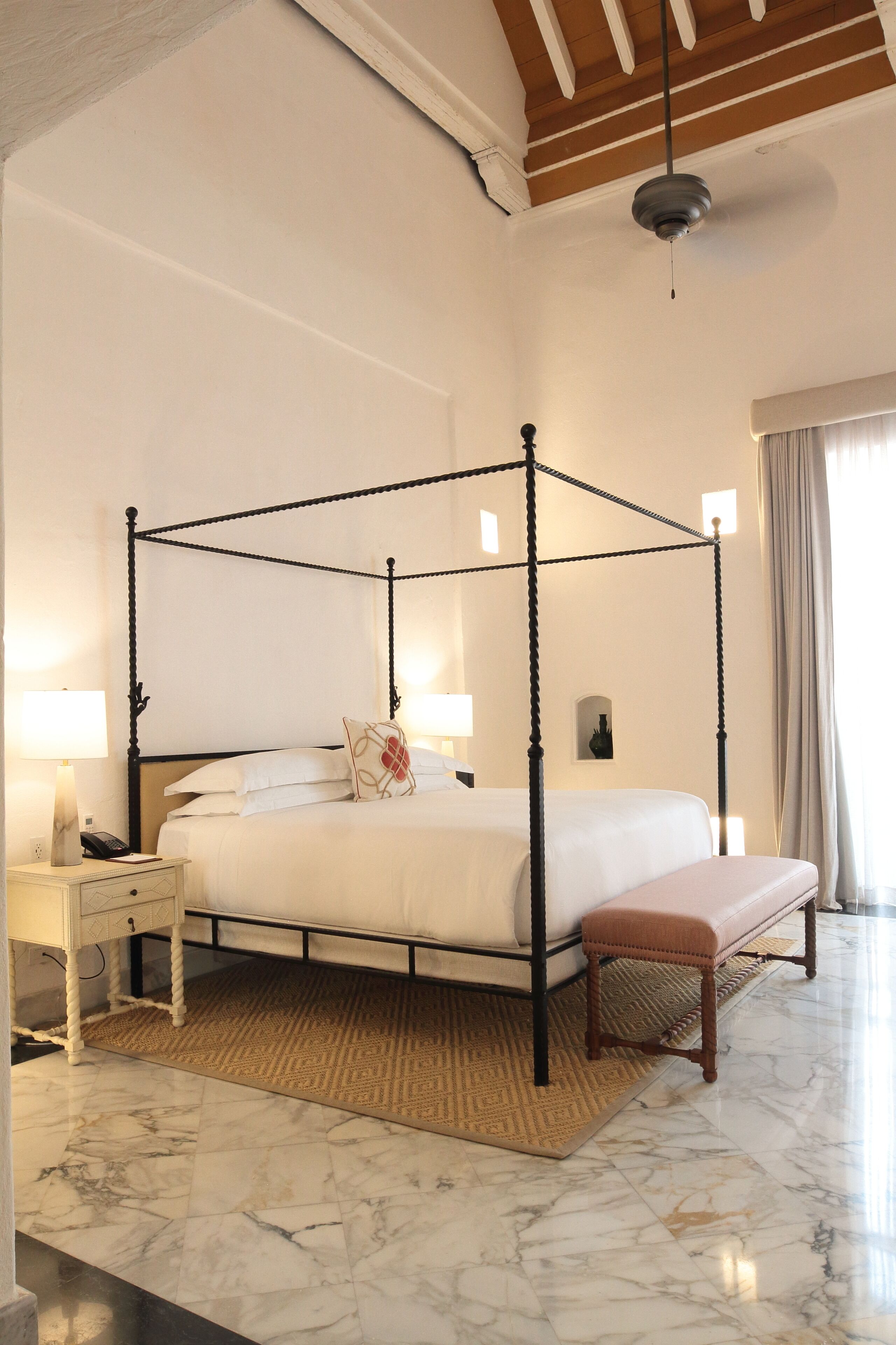 A four-poster bed with a wooden headboard, bedside tables with lamps, a bench, and a marble floor.