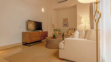 A living room with a flat-screen TV, a wooden cabinet, a white sofa, a round ottoman, and a floor lamp.