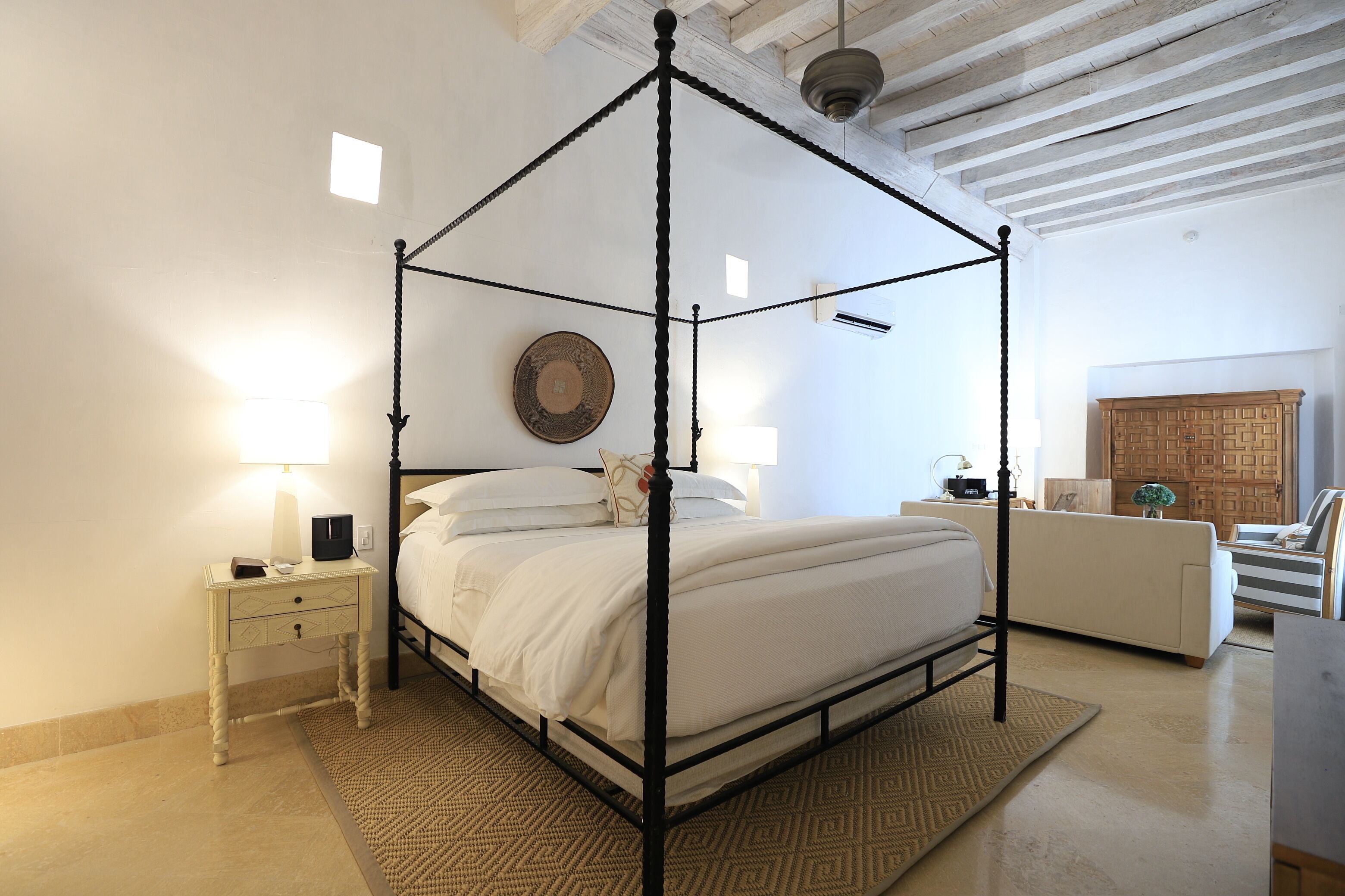 Una cama con dosel sobre un armazón de madera, una mesita de noche con lámpara y una alfombra estampada en el suelo.