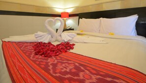 Quarto de hotel com uma cama, um cobreleito vermelho estampado, uma arrumação de toalhas em formato de coração e uma flor vermelha na cama.