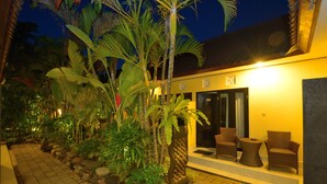 A well-lit outdoor area with a pathway lined with rocks and tropical plants, leading to a building with a reception desk and seating area. - Andari Legian (Legian)