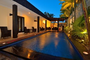 Uma área de piscina com cadeiras e um espaço de refeições, cercada por plantas e um muro de pedra.