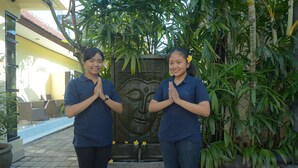 Two individuals standing in front of a decorative stone wall with a water feature, surrounded by lush greenery. - Andari Legian (Legian)