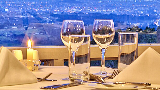 A table set for dining with a candle, three wine glasses, and cutlery, overlooking a cityscape at dusk. - Waitakere Resort & Spa (Auckland)