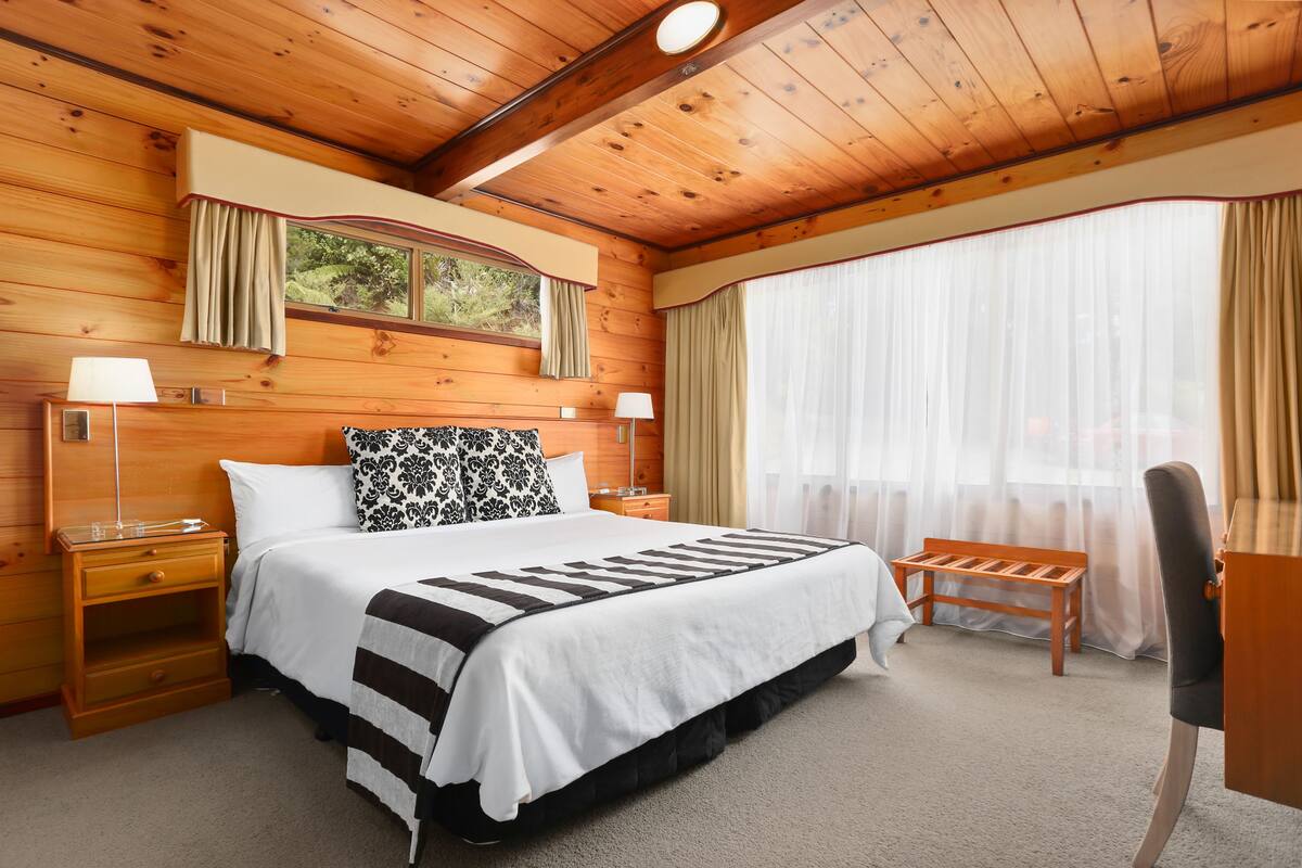 Une chambre à coucher aux murs lambrissés, avec un grand lit, deux tables de chevet avec des lampes, un banc et une fenêtre avec des rideaux.