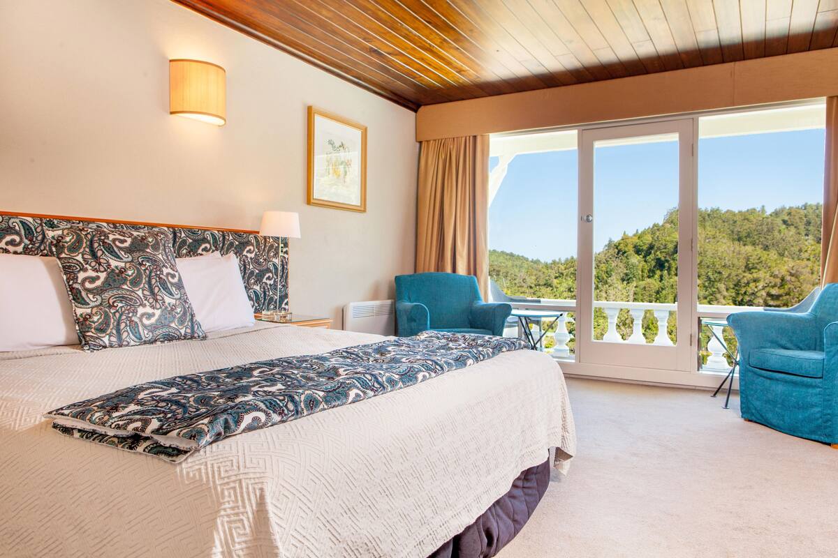 Une chambre d’hôtel avec un grand lit, deux fauteuils, un balcon donnant sur la forêt et un tableau au mur.
