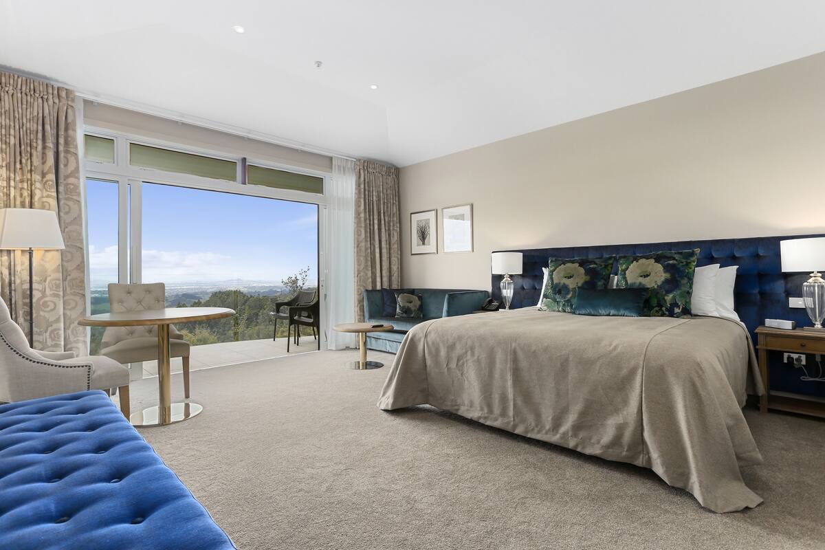 Une chambre d’hôtel avec un grand lit, un canapé, une petite table et une chaise. Une grande fenêtre offre une vue sur l’extérieur.