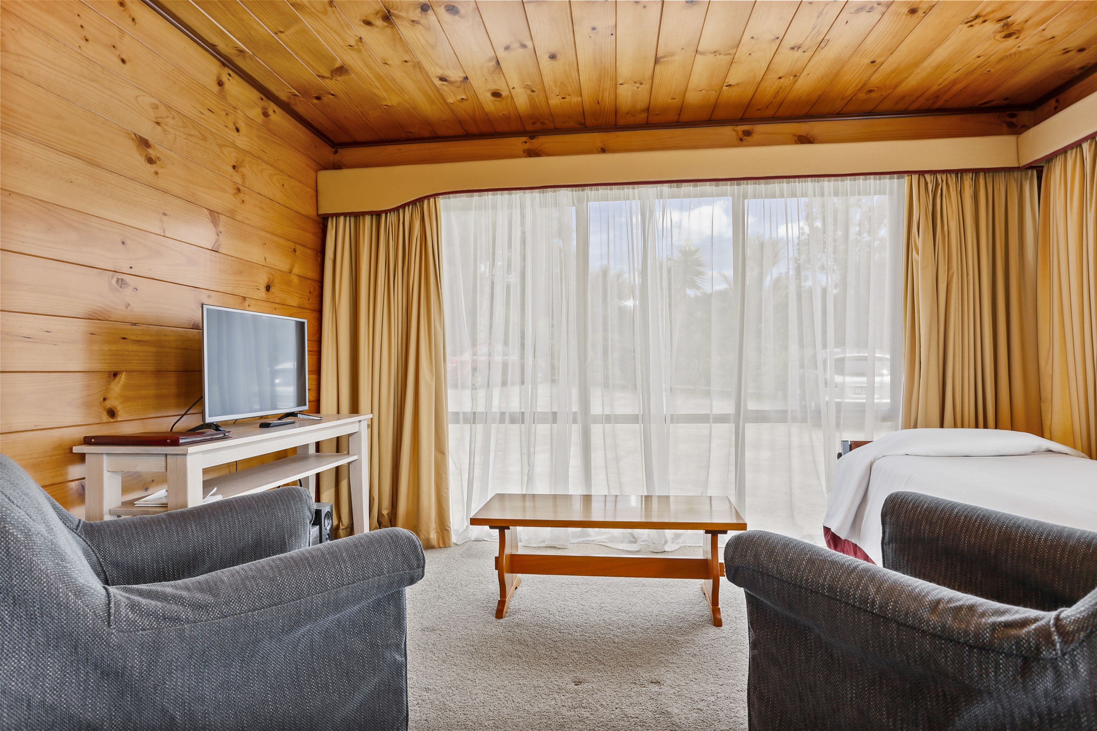 Una habitación con paredes de madera, un televisor sobre una mesita, dos sillones grises, una cama con ropa blanca y un amplio ventanal con cortinas traslúcidas.