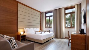 A modern hotel room with a large bed, a bedside table with a lamp, a TV, a wooden dresser, and a view of trees through the window. - Occidental Aurelia (Rome)