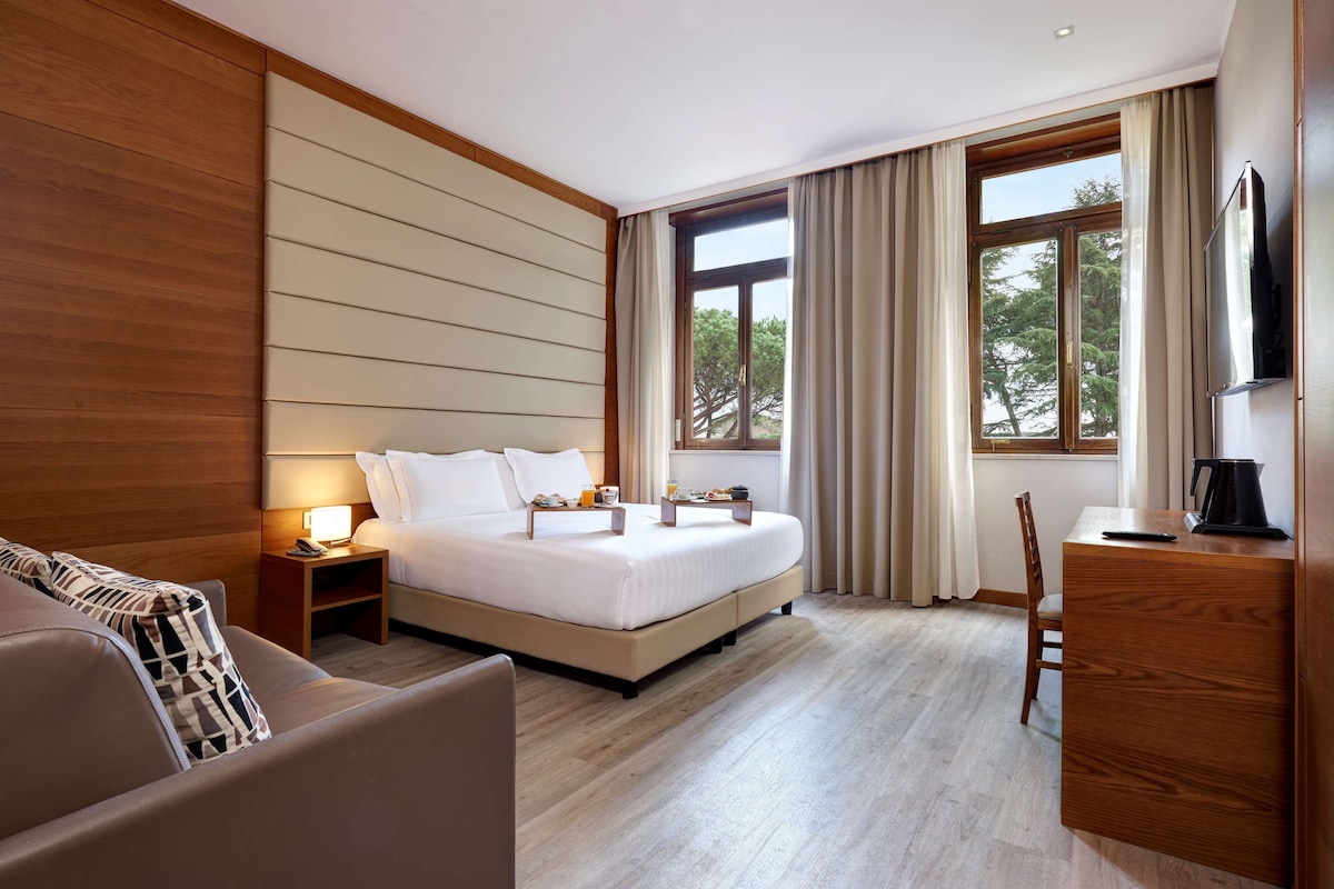 A modern hotel room with a large bed, a bedside table with a lamp, a TV, a wooden dresser, and a view of trees through the window.