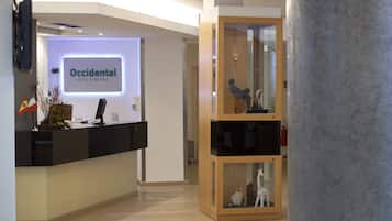 Un área de recepción con un cartel que dice “Occidental Hotel & Resorts”. Se observa un mostrador de recepción, un aparador de madera con figuritas y un diseño de interiores moderno.