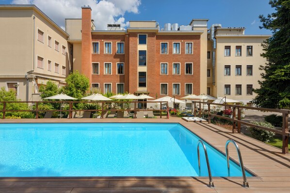 Una piscina con muelle de madera, rodeada de sombrillas y sillas de descanso, con edificios de varios pisos al fondo.